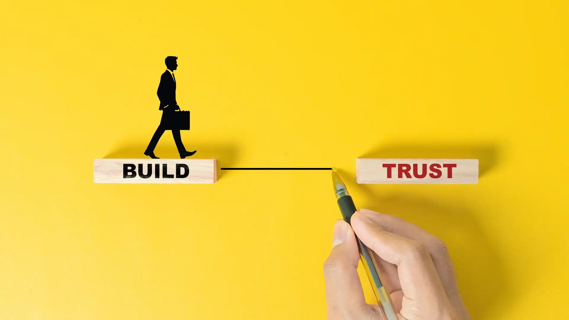 A businessman's hand building a stack of wooden blocks that spell out "BUILD TRUST" on a vibrant yellow table and background, symbolizing the creation of trust capital for Korean manufacturers entering the US market.